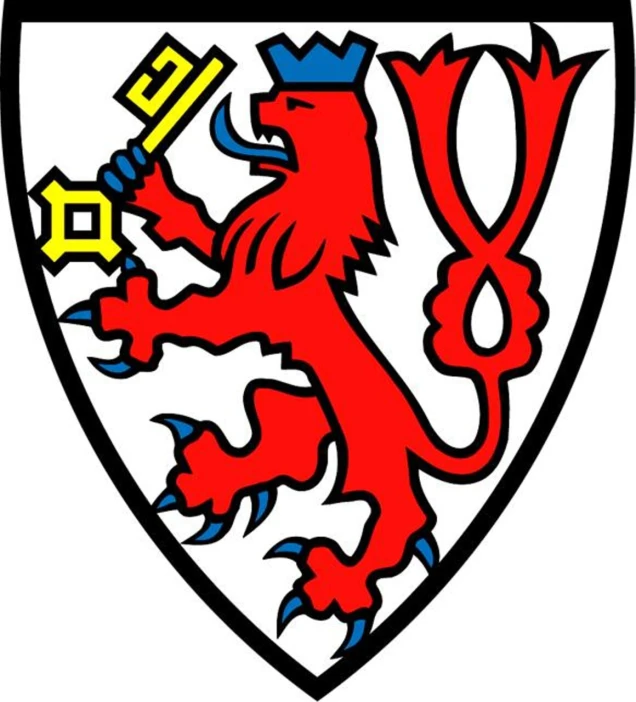 Wappen mit einem roten Löwen mit blauer Zunge und Krone, der einen goldenen Schlüssel hält, auf weißem Hintergrund mit schwarzer Umrandung.