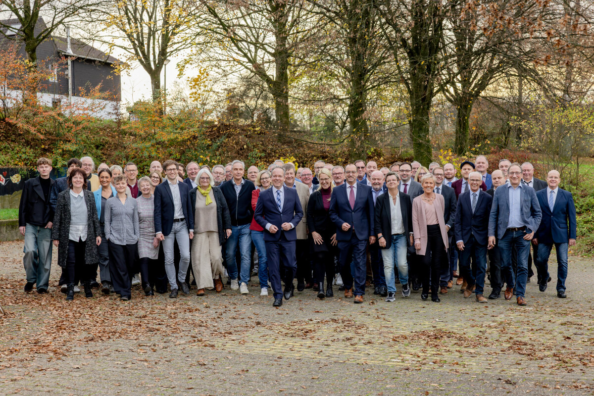 Eine große Gruppe von Menschen in herbstlicher Umgebung, einige in Businesskleidung, andere in Freizeitkleidung. Im Hintergrund sind Bäume und ein Gebäude sichtbar.