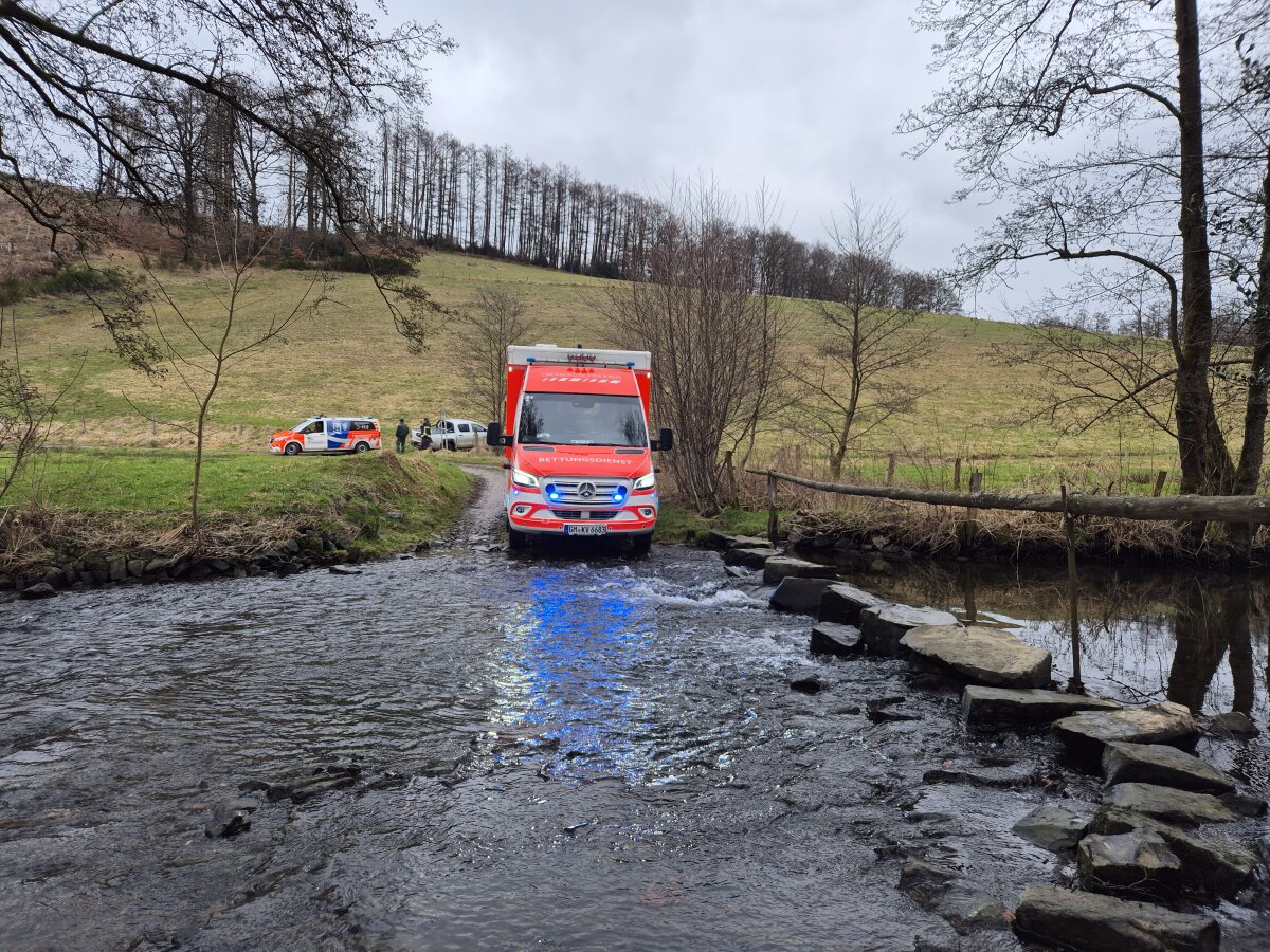 Ein rotes Rettungsdienstfahrzeug fährt durch einen flachen Bach. Im Hintergrund stehen ein weiteres Fahrzeug und mehrere Personen auf einer Wiese. Es ist bewölkt.