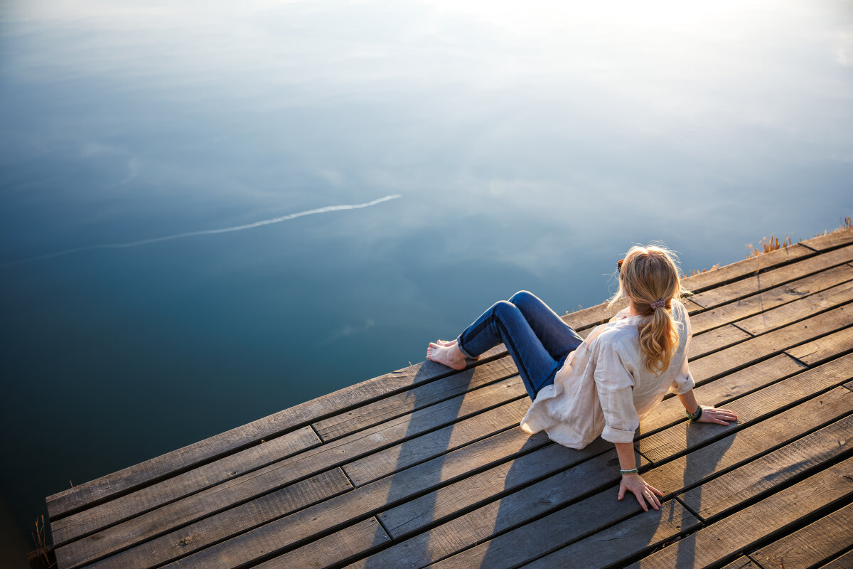Person mit heller Bluse und blonden Haaren sitzt auf einem hölzernen Steg am Wasser und blickt in die Ferne. Der Himmel spiegelt sich im ruhigen Wasser.