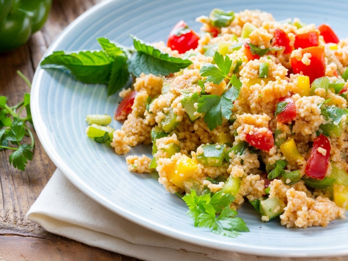 Ein Teller mit Couscous-Salat, garniert mit frischen Kräutern. Der Salat enthält bunte Paprikastücke, Gurken und Tomaten. Der Teller steht auf einem Holzuntergrund.