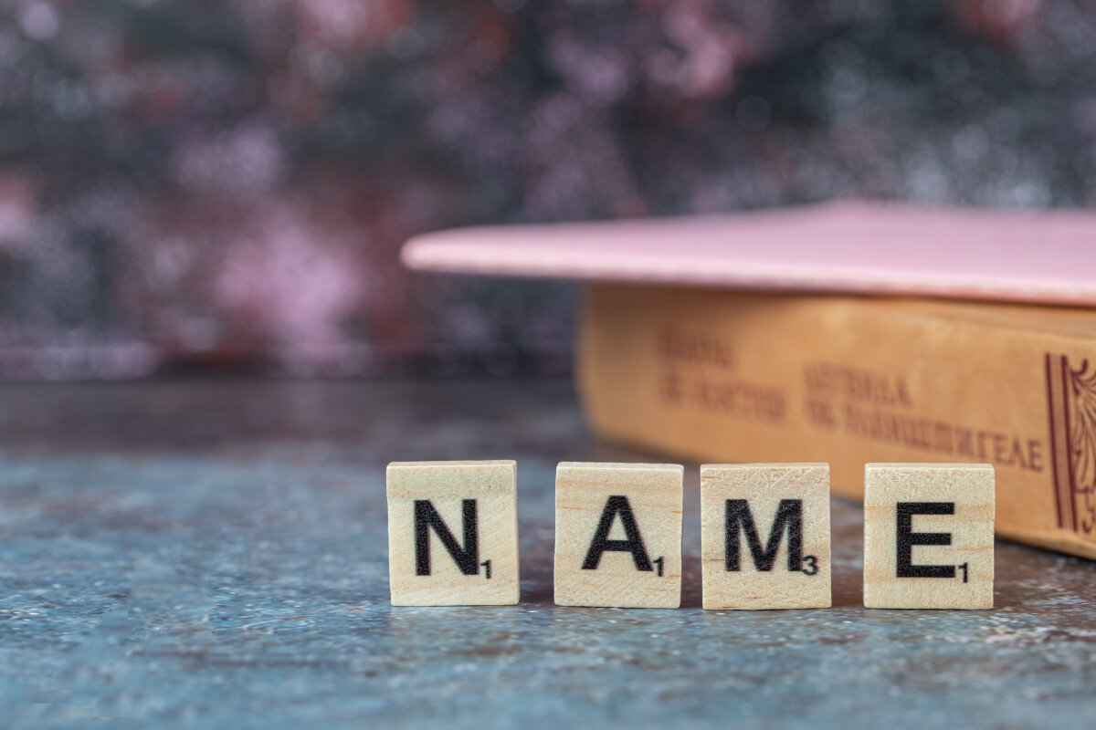 Vier Scrabble-Buchstabensteine mit den Buchstaben 'N', 'A', 'M' und 'E', die das Wort 'NAME' bilden, stehen auf einer Oberfläche. Im Hintergrund sind ein Buchrücken und eine verschwommene, bunte Fläche zu sehen.