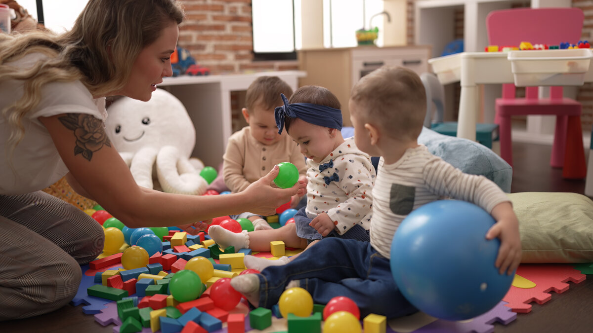 Eine Frau spielt mit drei Kleinkindern in einem Raum voller bunter Spielzeugbälle und Bausteine. Im Hintergrund sind Spielmöbel und ein großes Kuscheltier zu sehen.