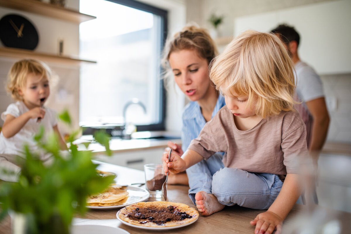 Eine Frau und zwei Kinder in einer Küche. Ein Kind sitzt auf der Arbeitsfläche und bestreicht einen Pfannkuchen, während das andere einen Löffel hält. Im Hintergrund ein Mann und eine Pflanze im Vordergrund.