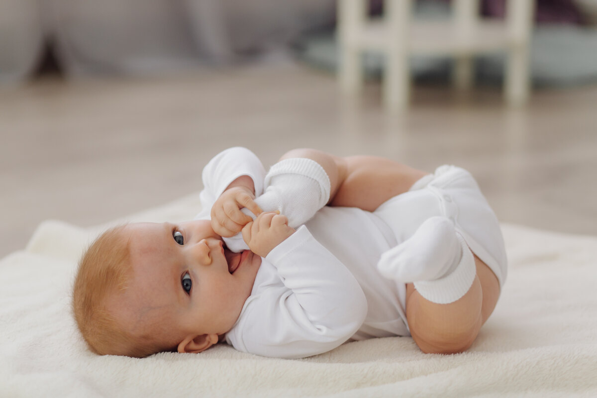 Ein Baby liegt auf dem Rücken auf einer weichen Decke, trägt einen weißen Strampler und weiße Söckchen und hält einen Fuß mit den Händen.