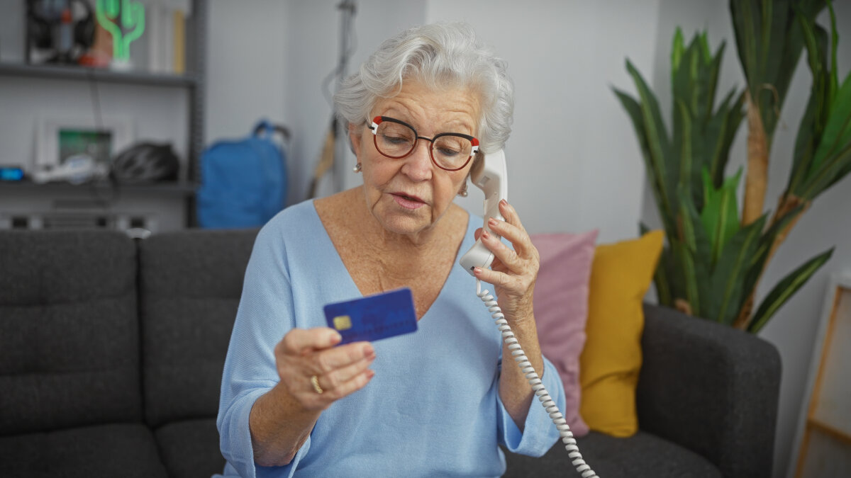 Eine ältere Frau mit Brille und grauen Haaren sitzt auf einem Sofa und telefoniert mit einem Festnetztelefon. In ihrer Hand hält sie eine blaue Kreditkarte. Im Hintergrund sind ein Regal, Kissen und eine Zimmerpflanze zu sehen.