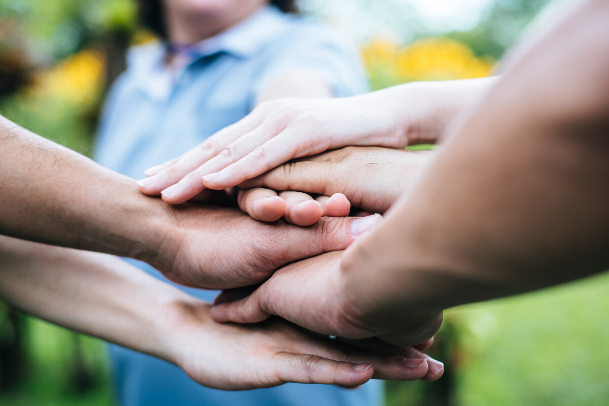 Mehrere Hände unterschiedlicher Personen, die übereinandergelegt sind, als Zeichen von Teamwork oder Zusammenhalt, mit unscharfem Hintergrund im Freien.