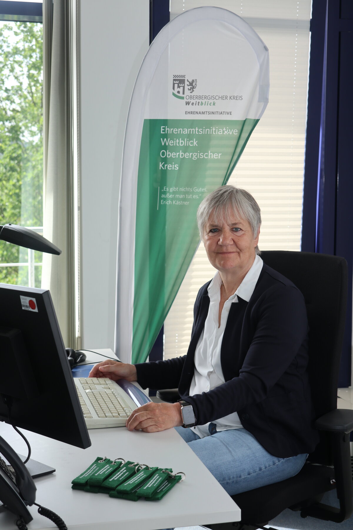 Eine Person sitzt an einem Schreibtisch mit Tastatur und Monitor. Im Hintergrund steht ein Banner mit der Aufschrift 'Ehrenamtsinitiative Weitblick Oberbergischer Kreis'.