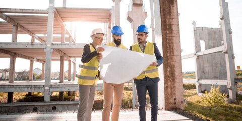 Drei Personen in gelben Warnwesten und Helmen stehen auf einer Baustelle und betrachten einen großen Bauplan. Im Hintergrund ist ein Rohbau aus Beton zu sehen.