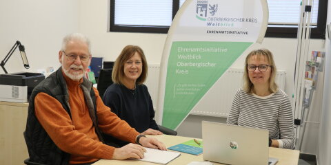 Drei Personen sitzen an einem Tisch in einem Büro. Ein Mann und zwei Frauen lächeln in die Kamera. Im Hintergrund ein Banner der Ehrenamtsinitiative Weitblick Oberbergischer Kreis sowie ein Fenster.