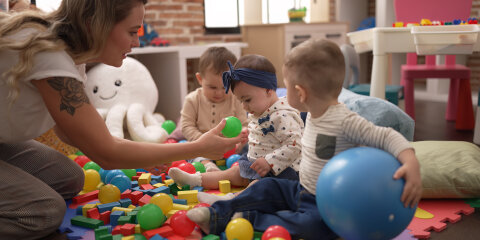 Eine Frau spielt mit drei Kleinkindern in einem Raum voller bunter Spielzeugbälle und Bausteine. Im Hintergrund sind Spielmöbel und ein großes Kuscheltier zu sehen.