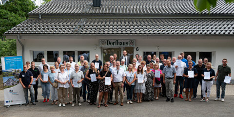 Eine Gruppe von Menschen posiert vor einem Gebäude mit der Aufschrift 'Dorhaus'. Einige Personen halten Urkunden in den Händen. Im Hintergrund sind Bäume sichtbar.