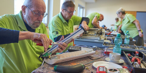 Mehrere Personen in grÃ¼nen Polohemden reparieren elektronische GerÃ¤te an einem Tisch in einem Innenraum. Auf dem Tisch liegen Werkzeuge und Bauteile.