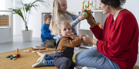 Eine Frau in rotem Oberteil spielt mit drei Kindern in einem hellen Raum. Spielzeug ist auf einem Teppich verteilt, und im Hintergrund stehen Regale mit weiteren Spielsachen.