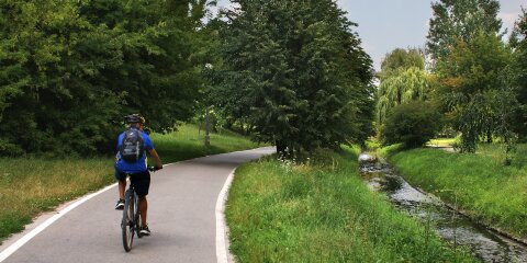 Ein Radfahrer mit Helm und Rucksack fährt auf einem asphaltierten Weg durch eine grüne Landschaft. Rechts neben dem Weg fließt ein schmaler Bach.