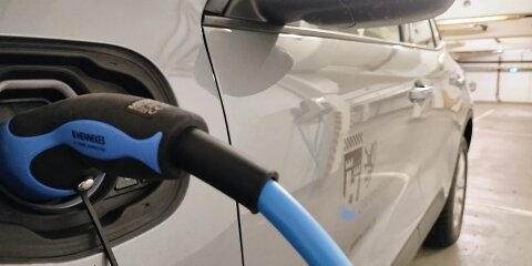 Ein Elektroauto wird in einer Tiefgarage mit einem blauen Ladekabel aufgeladen. Der Ladeanschluss und ein Seitenspiegel des Autos sind sichtbar.
