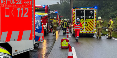 Einsatzfahrzeuge und Rettungskräfte auf einer nassen Straße, darunter Rettungswagen, Feuerwehrfahrzeuge und Personen in Einsatzkleidung. Verkehrshütchen sichern den Bereich ab, im Hintergrund ist eine Brücke sichtbar.