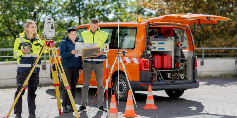 Drei Personen in Sicherheitskleidung arbeiten mit Vermessungsgeräten vor einem orangefarbenen Fahrzeug mit geöffnetem Kofferraum auf einem Parkplatz.