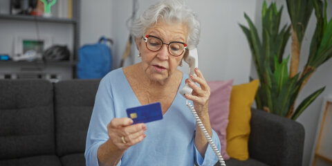 Eine Ã¤ltere Frau mit Brille und grauen Haaren sitzt auf einem Sofa und telefoniert mit einem Festnetztelefon. In ihrer Hand hÃ¤lt sie eine blaue Kreditkarte. Im Hintergrund sind ein Regal, Kissen und eine Zimmerpflanze zu sehen.