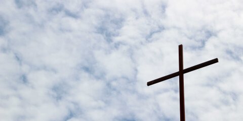 Ein einfaches, dunkles Kreuz auf der Spitze eines Gebäudes vor einem bewölkten Himmel.