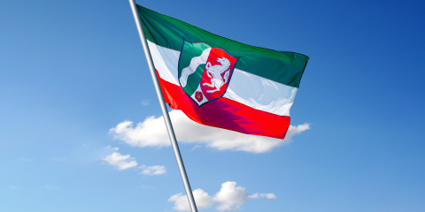 Flagge mit horizontalen Streifen in Grün, Weiß und Rot, darauf ein Wappen mit einem weißen Pferd, einer roten Blume und einem geschwungenen Element, vor blauem Himmel mit Wolken.
