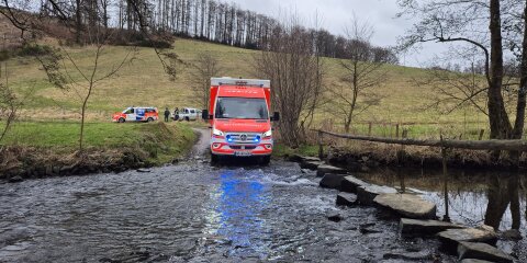 Ein rotes Rettungsdienstfahrzeug fährt durch einen flachen Bach. Im Hintergrund stehen ein weiteres Fahrzeug und mehrere Personen auf einer Wiese. Es ist bewölkt.