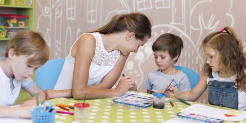 Eine Frau sitzt mit drei Kindern an einem Tisch, der mit einer grünen Tischdecke mit weißen Punkten bedeckt ist. Sie malen mit Wasserfarben und Stiften. Im Hintergrund ist eine Wand mit gemalten weißen Haus- und Baumumrissen zu sehen.