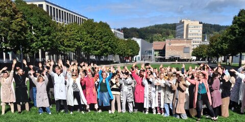 Eine große Gruppe von Menschen steht auf einer Wiese, viele tragen Bademäntel und heben die Arme in die Luft. Im Hintergrund sind Gebäude und Bäume zu sehen.
