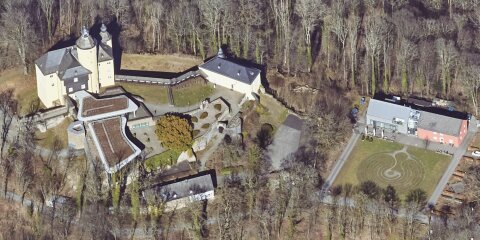 Schrägluftbild einer Schlossanlage mit gelbem Hauptgebäude, Nebengebäuden, Gartenanlagen und einem modernen Anbau. Im Vordergrund eine Wiese mit Labyrinthmuster.