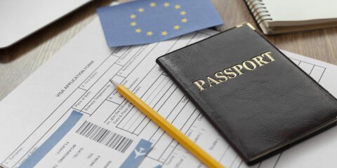 Ein brauner Reisepass, ein gelber Bleistift, ein Visa-Antragsformular, Flugtickets und eine EU-Flagge liegen auf einem Holztisch. Im Hintergrund ein Laptop und ein Notizbuch.