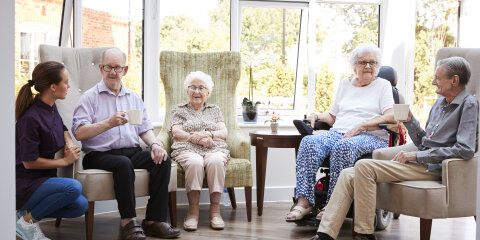 Male And Female Residents Sitting In Chairs And Talking With Carer In Lounge Of Retirement Home