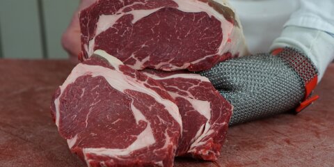 Zwei rohe Steakstücke mit sichtbarer Fettmarmorierung liegen auf einem Schneidebrett. Eine Hand mit einem Kettenhandschuh hält die Steaks.