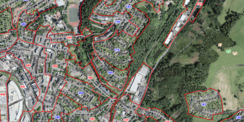 Luftbild von Gummersbach mit markierten Bodenrichtwertzonen, die durch rote Linien getrennt sind. Häuser, Straßen und Grünflächen sind sichtbar. Zahlen in verschiedenen Farben und Größen zeigen Bodenrichtwerte an..
