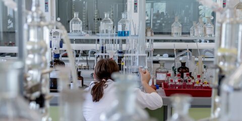 Eine Person in einem weißen Laborkittel arbeitet in einem Labor, umgeben von chemischen Glasgefäßen, Reagenzflaschen und Pipetten auf einem Labortisch.