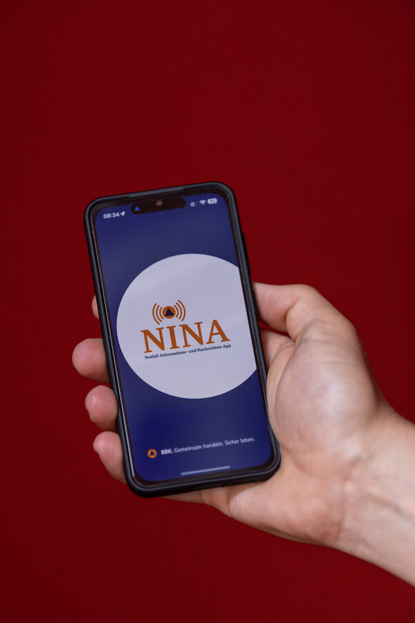 Eine Hand hält ein Smartphone, auf dessen Bildschirm die Startseite der NINA-App angezeigt wird. Der Hintergrund ist rot.