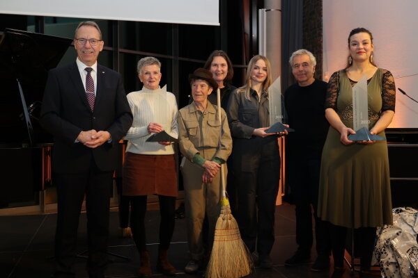 Eine Gruppe von sieben Personen steht vor einer Bühne, darunter vier Frauen mit Auszeichnungen in der Hand und eine Person mit Besen. Ein Mann trägt einen Anzug.