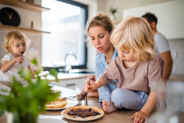 Eine Frau und zwei Kinder in einer Küche. Ein Kind sitzt auf der Arbeitsfläche und bestreicht einen Pfannkuchen, während das andere einen Löffel hält. Im Hintergrund ein Mann und eine Pflanze im Vordergrund.