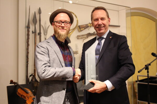 Zwei Männer stehen nebeneinander, schütteln sich die Hand und halten eine Auszeichnung. Im Hintergrund sind historische Waffen und ein Geigenbogen ausgestellt.
