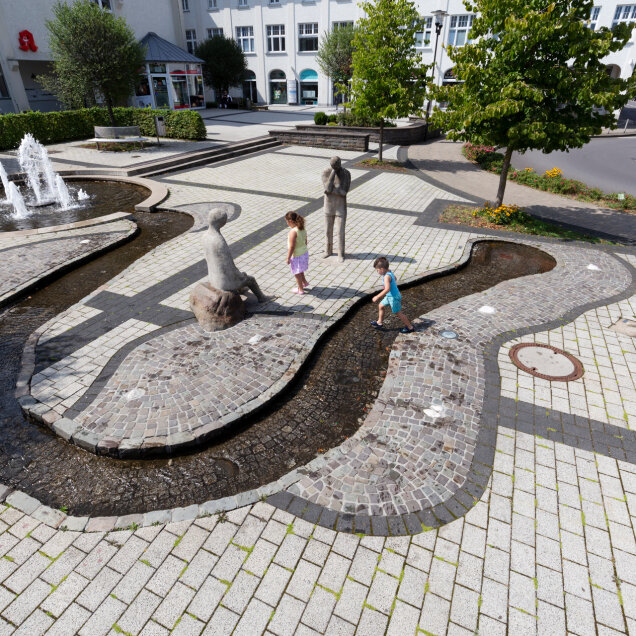 Ein öffentlicher Platz mit einem geschwungenen Wasserlauf, mehreren Brunnen und Figuren aus Stein. Zwei Kinder spielen im Wasser. Gebäude und Bäume im Hintergrund.