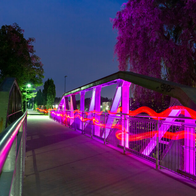 Eine Brücke bei Nacht mit leuchtenden pinken und roten Lichteffekten entlang des Geländers. Im Hintergrund sind Bäume und ein dunkler Himmel sichtbar.