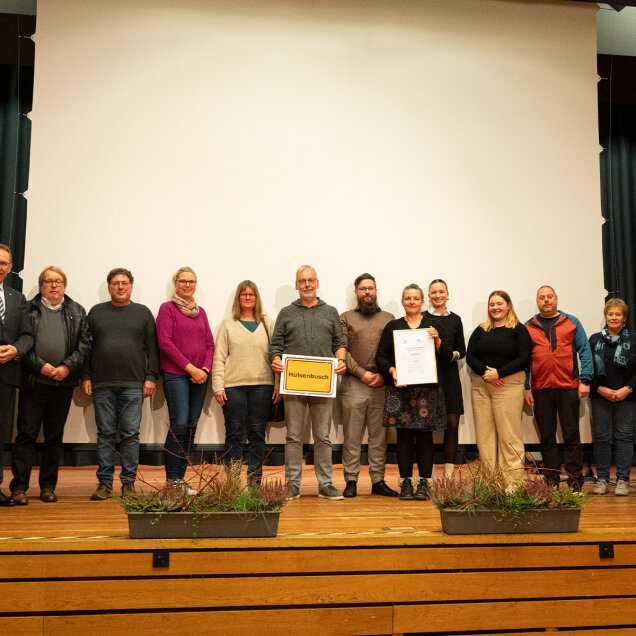 Eine Gruppe von Personen steht auf einer Bühne vor einer weißen Leinwand. Einige halten ein Schild und eine Urkunde. Vorne auf der Bühne stehen Blumenkästen.