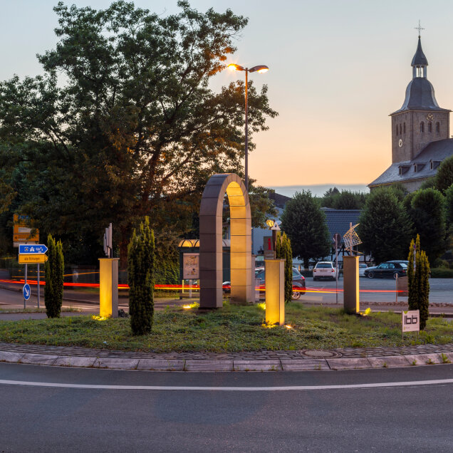 Beleuchteter Kreisverkehr mit einer modernen Skulptur im Zentrum, umgeben von Bäumen und parkenden Autos. Im Hintergrund eine Kirche bei Sonnenuntergang.