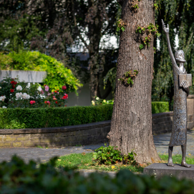 Bronzestatue einer stehenden Person, die einen Arm nach oben hebt und ein Buch hält. Die Statue steht neben einem Baum in einer gepflegten Gartenanlage mit Blumen.
