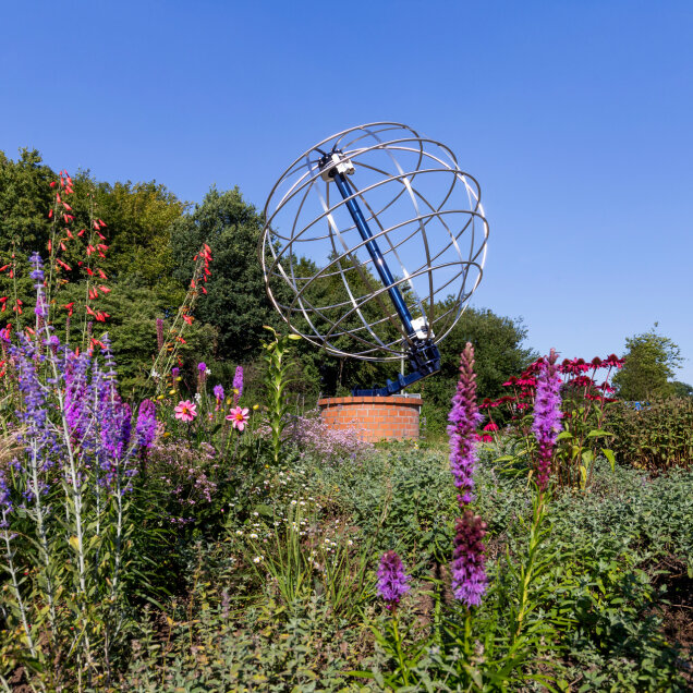Eine metallische Globuskonstruktion auf einem gemauerten Sockel, umgeben von blühenden Pflanzen in verschiedenen Farben, darunter lila, rot und rosa. Im Hintergrund sind grüne Bäume und ein blauer Himmel sichtbar.