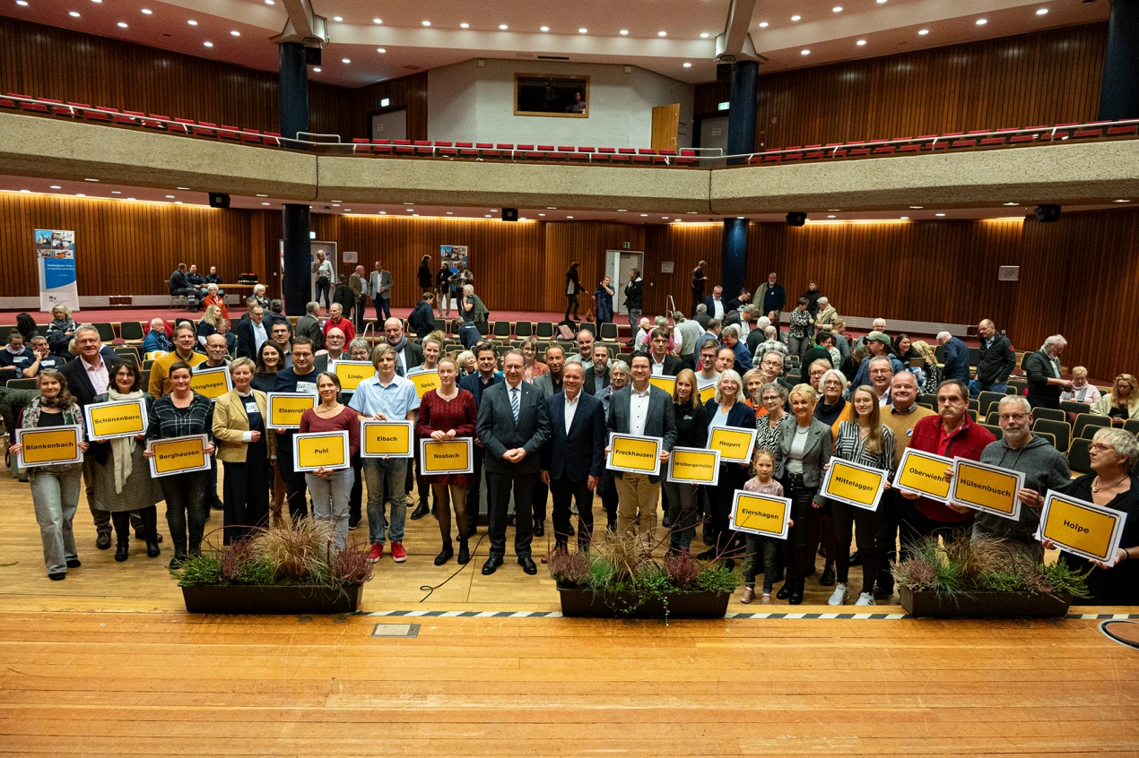Eine größere Gruppe von Menschen steht in einem Saal mit Holzboden und Holzwänden. Einige Personen halten Schilder mit Ortsnamen. Im Hintergrund stehen weitere Personen und Stühle.