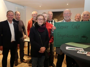 Eine Gruppe von Personen steht in einem Raum und lächelt. Eine Person hält ein grünes Schild mit der Aufschrift 'Repair Café Engelskirchen' und einer gezeichneten Kaffeetasse.