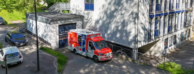 Ein Rettungswagen steht vor einem Gebäude mit der Beschriftung 'Rettungsdienst Ausfahrt frei'. Im Vordergrund sind parkende Autos und eine grüne Umgebung sichtbar.