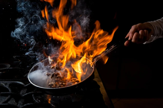 Eine Hand hält eine Pfanne mit brennenden Zutaten, Flammen und Rauch steigen auf, dunkler Hintergrund und Gaskochfeld sichtbar.