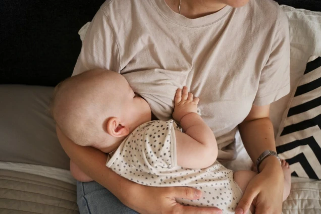 Eine Person stillt ein Baby, das ein gepunktetes Oberteil trägt. Die Person trägt ein helles T-Shirt und hält das Baby im Arm. Im Hintergrund ist ein Kissen zu sehen.