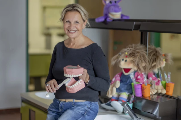 Eine lächelnde Frau sitzt auf einem Waschbecken und hält ein Modell eines Gebisses mit einer Zahnbürste. Im Hintergrund sind bunte Puppen und Zahnbürsten zu sehen.